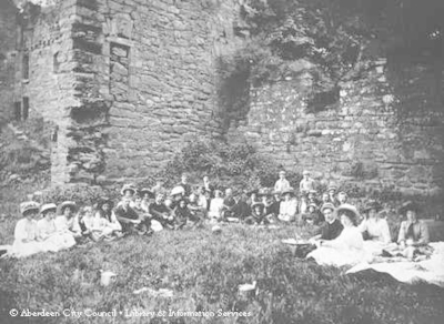 Picnic scene of mostly ladies and children in the grounds of a ruined castle