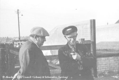 Two men outside in work clothes and caps, one holding a pipe