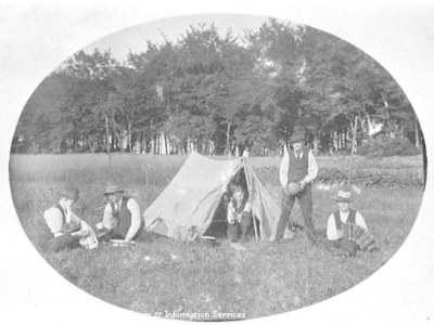 Portrait of young men relaxing outside tent. One plays an accordion.